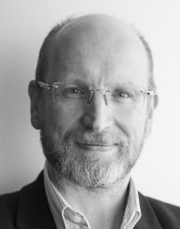 A black and white headshot of David Knott, wearing glasses, a light open-collared shirt and a dark jacket